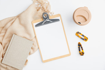 Elegant home office desk with blank paper clipboard, beige blanket, notebook, cup of coffee and barrettes on white background. Flat lay, top view. Minimalistic feminine workspace concept.