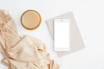 Flat lay mobile phone mockup with blank white screen, paper notebook, cup of coffee and beige blanket on white table. Top view modern home office desk.