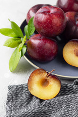 Whole purple plums and slices with leaves and knife on ceramic plate, light grey concrete background.
