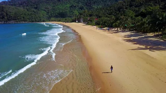 Directional high aerial drone flight over waves crashing at colorful empty deserted paradise beach Tioman Island Malaysia. Yellow sand with forest, palm trees and mountains in the background.
