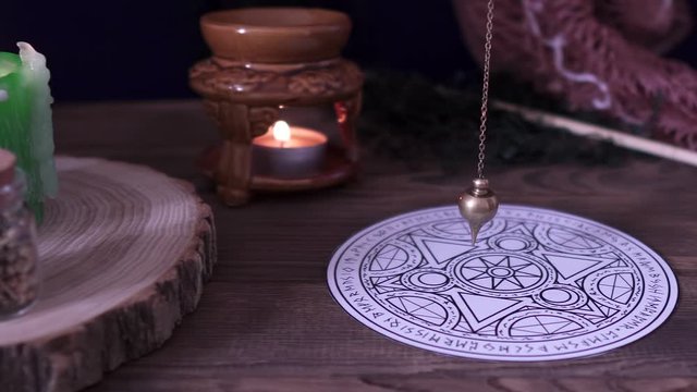 Witchcraft and wizardry. The sorcerer uses the esoteric pendulum for Tarot and the occult circle for divination next to burning candle and magical potions and subjects. Low light, close-up