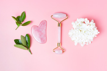 Facial roller and gua sha scraper with green leaves on pink background, concept of facial massage