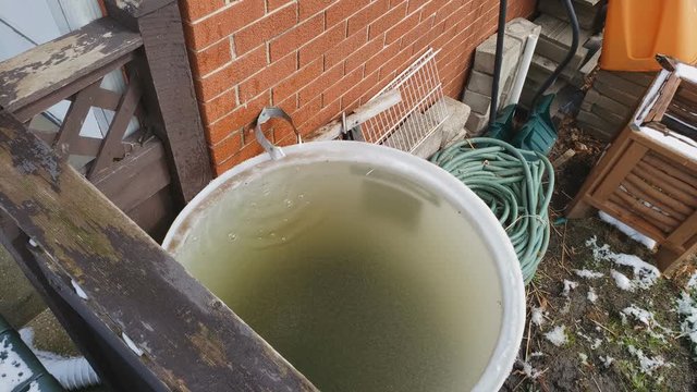 Top view of water dripping from melting snow from gutter into large barrel full of stored freezing water for future gardening.