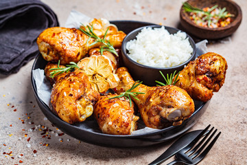 Baked spicy chicken legs with rice, garlic and herbs in black plate, dark background.