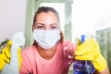 Fototapeta na wymiar Woman with protective glove and facial mask doing housework, housewife portrait. She sprinkling disinfectant and cleaning the glass. Stay safe.