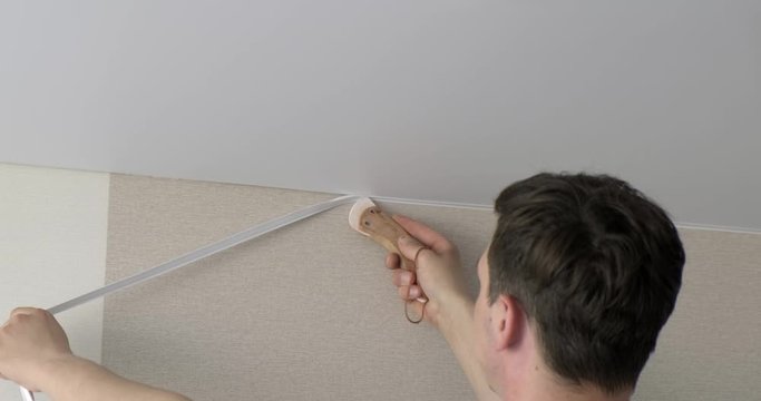 Male builder with special spatula straightens edge of ceiling into seam connection, fixing stretch ceiling to wall, decorative seam, suspended ceiling, construction and repair work in apartment