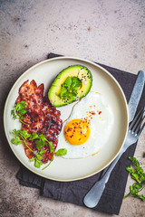 Keto breakfast - fried egg, avocado and fried bacon in white plate, top view. Keto diet concept.