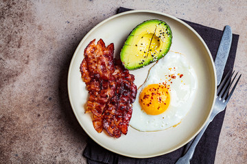 Keto breakfast - fried egg, avocado and fried bacon in white plate, top view. Keto diet concept.