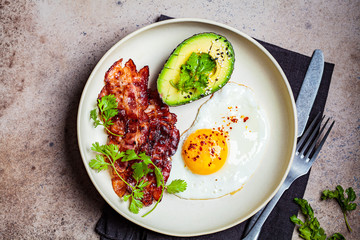 Keto breakfast - fried egg, avocado and fried bacon in white plate, top view. Keto diet concept.