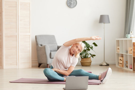 Senior woman sitting on the floor on exercise mat and stretching her hands while watching sports training on laptop at home