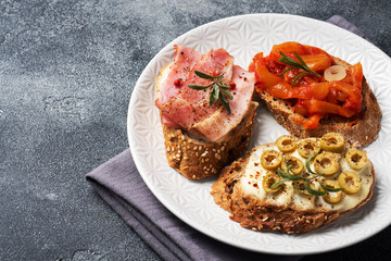 Wholegrain bread sandwiches with cream cheese, bacon and olives canned peppers with tomato on a plate. Dark concrete background with Copy space.