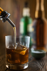 In the bar, whiskey is poured into a glass with ice, in the background there are bottles on a wooden table of the bar counter, shallow depth of field, selective focus. The concept of alcoholic drinks