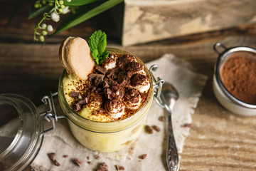 Tiramisu in a jar, homemade Italian dessert with mascarpone cheese, eggs, chocolate, cacao on rustic wooden table with flowers. Natural sunny light in the kitchen. Selective focus, copy space.