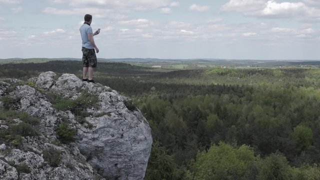 The man walking on top of rock, stops and starts taking pictures. A man on a cliff takes a photo with his mobile phone on sunny day. one person 29.97fps