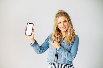 Take a look at this. Attractive young woman points finger to a blank phone screen, white wall on...