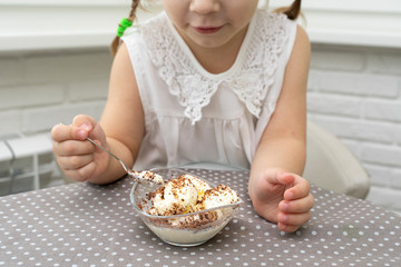 little girl in a white blouse happily eats ice cream from a snail at a table in the kitchen