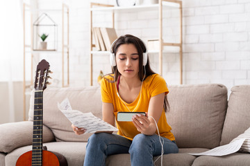 Millennial girl in headphones learning to play guitar by watching online video tutorial on mobile phone, indoors
