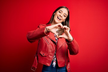 Young beautiful brunette woman wearing casual jacket standing over red background smiling in love doing heart symbol shape with hands. Romantic concept.