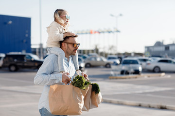 Caucasian father shopping in grocery store with baby daughter. Dad buying fresh vegetables. A happy father with a small daughter is walking in the parking lot holding paper bags in his hands