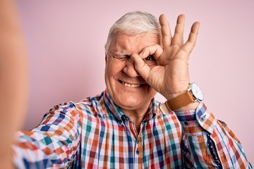 Senior handsome hoary man wearing casual shirt making selfie by the camera with happy face smiling doing ok sign with hand on eye looking through fingers