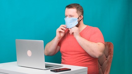 A bearded man in a pink T-shirt puts on a protective medical mask for working at a laptop at home on a green background. Quarantine concept