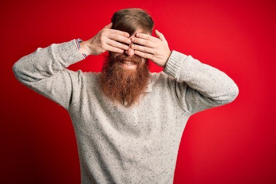 Handsome Irish redhead man with beard wearing casual sweater and glasses over red background covering eyes with hands smiling cheerful and funny. Blind concept.