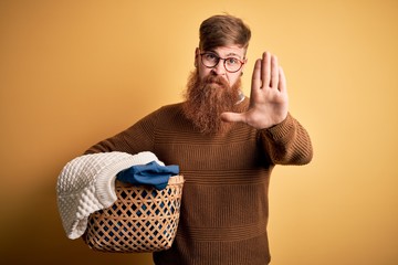Redhead Irish man with beard doing housework laundry over yellow background with open hand doing...