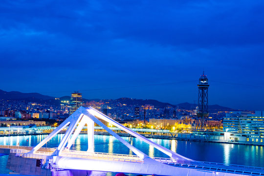Barcelona, Spain. Porta d’Europa bascule mobile bridge lit with night lights. Torre Jaume I aerial lift pylon steel truss tower, city skyline architecture and mountains behind.