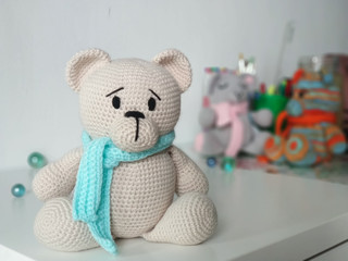 crochet bear beige in blue scarf on background of other toys on light background. Knitted soft toy Teddy