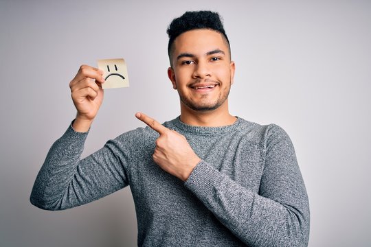 Handsome man holding reminder paper with sad emotion face emoji over white background very happy pointing with hand and finger