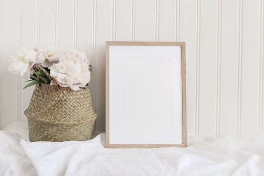 Beige blank wooden picture frame mockup. Artistic table still life composition with pink peony flowers in straw basket. White wooden wall background. Empty copyspace, rustic design.