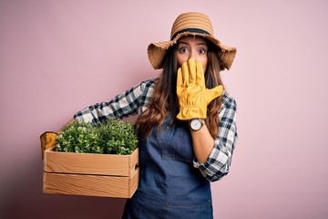 Young beautiful brunette farmer woman wearing apron and hat holding box with plants cover mouth with hand shocked with shame for mistake, expression of fear, scared in silence, secret concept