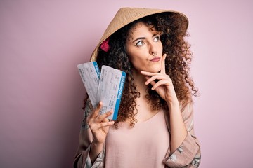 Young beautiful woman with curly hair and piercing wearing asian hat holding boarding pass serious face thinking about question, very confused idea