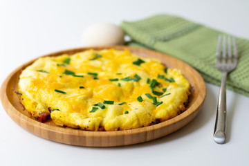 Fluffy omelette from whipped eggs with cheddar cheese and green onion scallions sprinkled on it in a wooden plate. It is surrounded by scallions, an egg, basil leaves, cheese, a green napkin, a fork