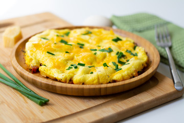 Fluffy omelette from whipped eggs with cheddar cheese and green onion scallions sprinkled on it in a wooden plate. It is surrounded by scallions, an egg, basil leaves, cheese, a green napkin, a fork
