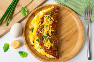 Fluffy folded egg omelette with tomato, red bell pepper, garlic, scallions and cheddar cheese in a wooden plate. In the background are an egg, basil leaves, scallions, cheese, napkin and a fork