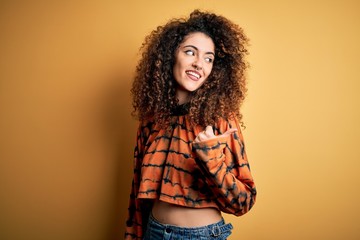 Young beautiful brunette woman with curly hair and piercing wearing casual t-shirt smiling with happy face looking and pointing to the side with thumb up.