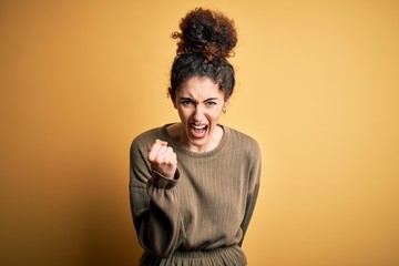 Young beautiful brunette woman with curly hair and piercing wearing casual dress angry and mad raising fist frustrated and furious while shouting with anger. Rage and aggressive concept.