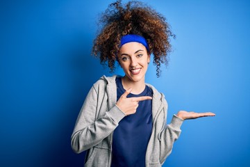 Young beautiful sportswoman with curly hair and piercing standing wearing sportswear amazed and smiling to the camera while presenting with hand and pointing with finger.