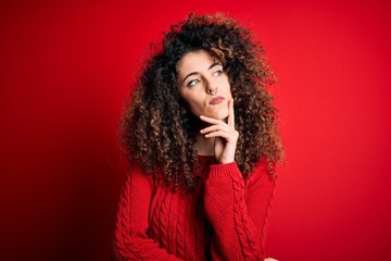Young beautiful woman with curly hair and piercing wearing casual red sweater with hand on chin thinking about question, pensive expression. Smiling with thoughtful face. Doubt concept.