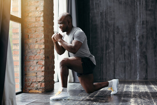 Muscular african american trainer in tight t-shirt doing an exercise lunging forward while holding hands at chest