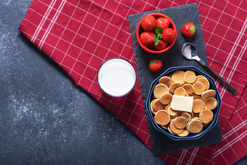 mini cereal pancakes in blue bowl with buttering toast, strawberries, spoon, glass of milk on grey background, rural Breakfast on red kitchen towel