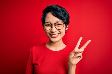 Young beautiful asian girl wearing casual t-shirt and glasses over isolated red background showing and pointing up with fingers number two while smiling confident and happy.
