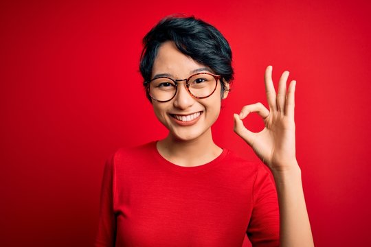 Young beautiful asian girl wearing casual t-shirt and glasses over isolated red background smiling positive doing ok sign with hand and fingers. Successful expression.
