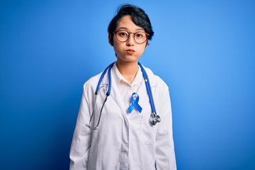 Young beautiful asian doctor girl wearing stethoscope and coat with blue cancer ribbon puffing cheeks with funny face. Mouth inflated with air, crazy expression.