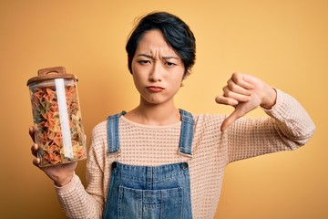 Young beautiful asian girl holding jar with italian dry pasta over isolated yellow background with...