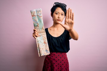 Young beautiful asian tourist girl on vacation holding city map over isolated pink background with open hand doing stop sign with serious and confident expression, defense gesture