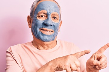 Senior man with grey hair wearing mud mask smiling and looking at the camera pointing with two hands and fingers to the side.