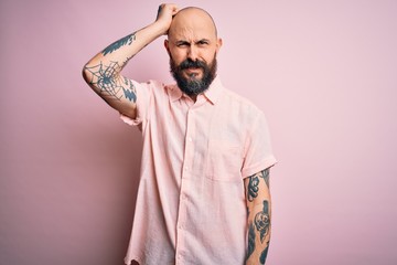 Handsome bald man with beard and tattoo wearing casual shirt over isolated pink background confuse and wonder about question. Uncertain with doubt, thinking with hand on head. Pensive concept.