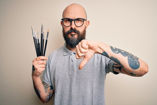 Handsome bald artist man with beard and tattoo painting using painter brushes with angry face, negative sign showing dislike with thumbs down, rejection concept